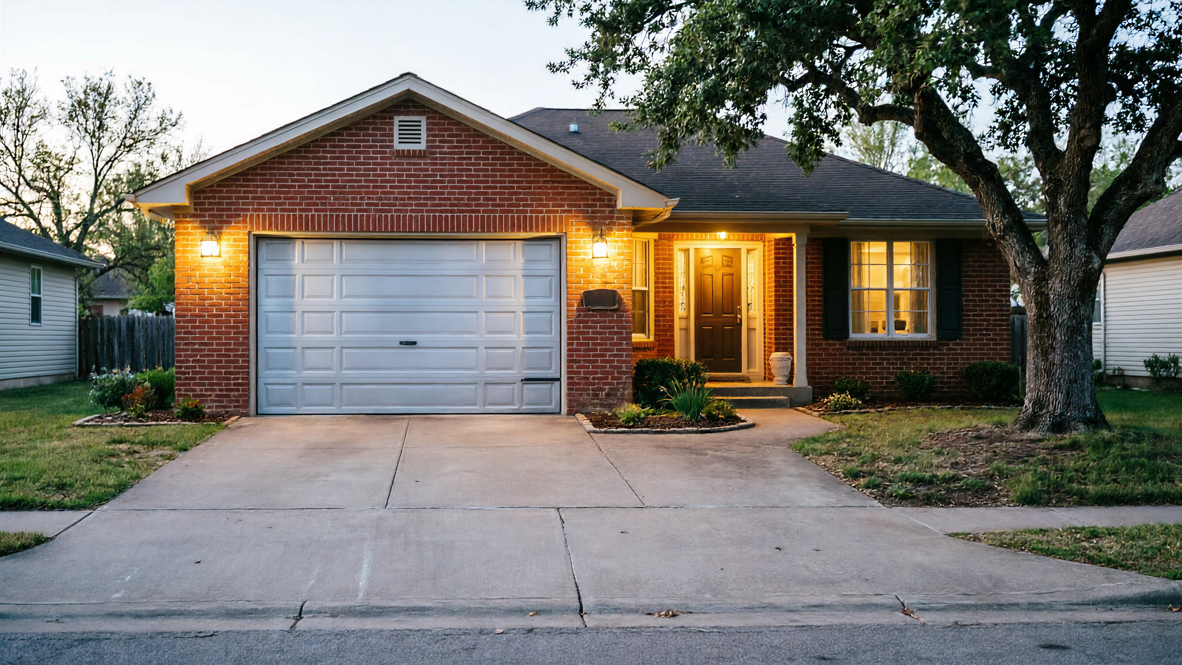 A brick house with a white garage door, a front door, and a large tree.